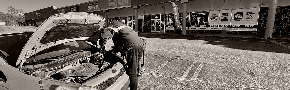A parts-store employee changing a car battery in the parking lot outside the shop
