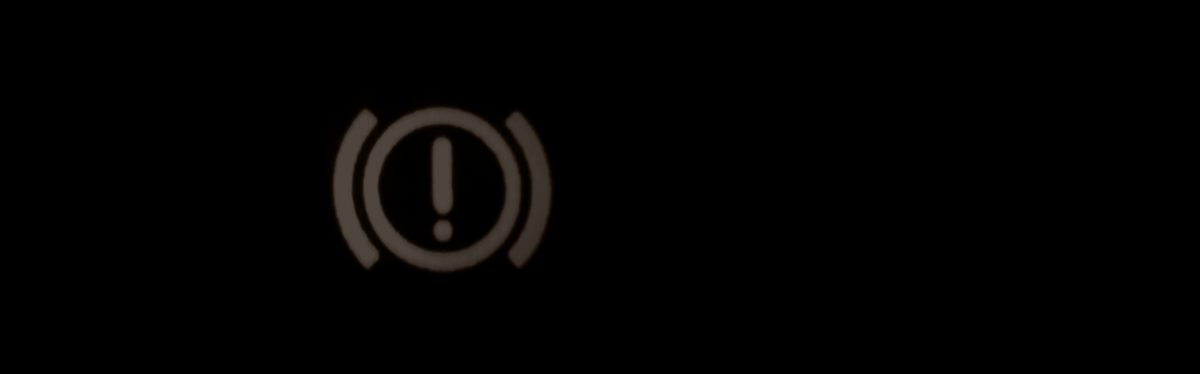 A red brake warning lamp, an exclamation mark in a circle, lit on a dark instrument cluster