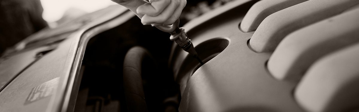 A hand pulls the oil dipstick out of a car engine under a raised hood to check the oil level.