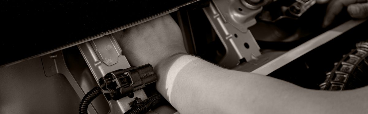 A two-pin electrical connector pulled apart on a corrugated wiring loom in an engine bay, with a hand reaching in behind it