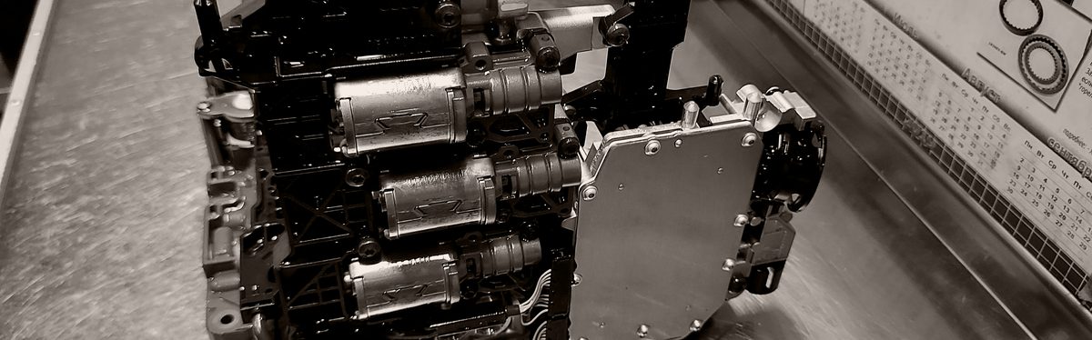 A transmission valve body on a workbench, its shift solenoids in a row beside the control module bolted to its side