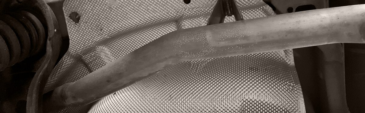 The underside of a car: a dimpled metal heat shield with the exhaust pipe across it and its rubber hanger at the top