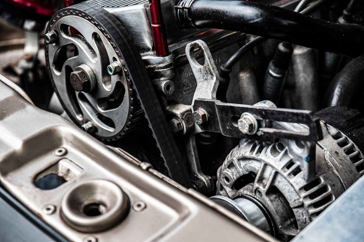 An alternator low on an engine, its finned casing and pulley turned by the ribbed belt running up to the sprocket above