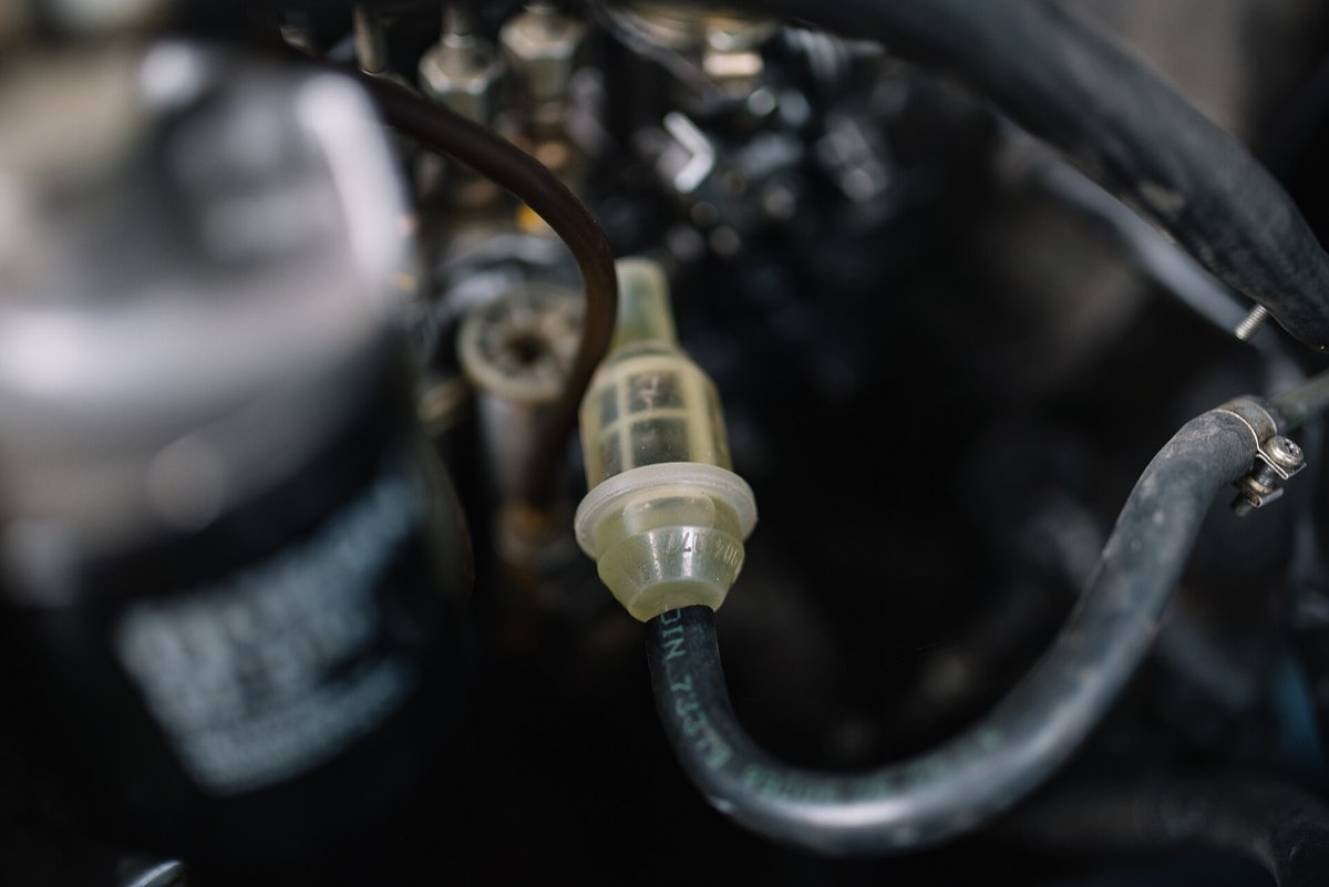A translucent inline fuel filter spliced into a rubber fuel line in an engine bay, with the fuel pump and hoses behind it