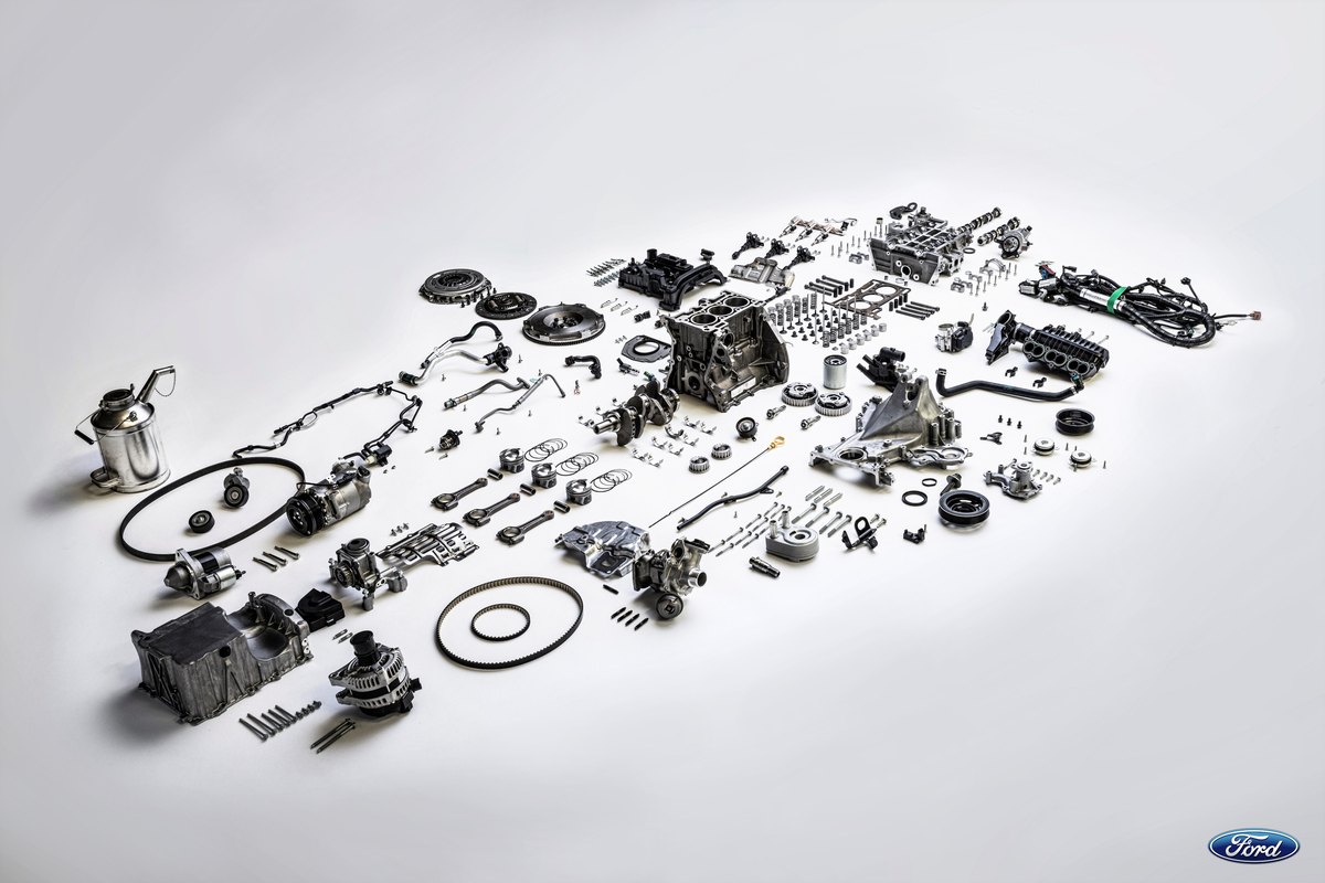 Every part of a Ford 1.0 EcoBoost engine laid out on a white floor, with a toothed rubber belt among them