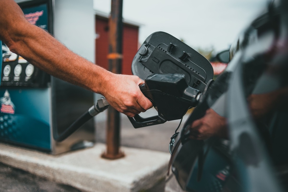 Hand holding a fuel nozzle in an open filler neck at a gas station, filler door swung back