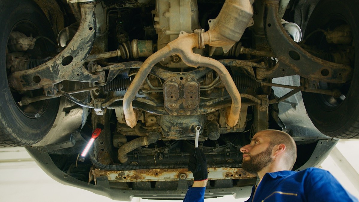 A mechanic under a lifted car looks up at the exhaust, where the converter sits between the manifold and the muffler.