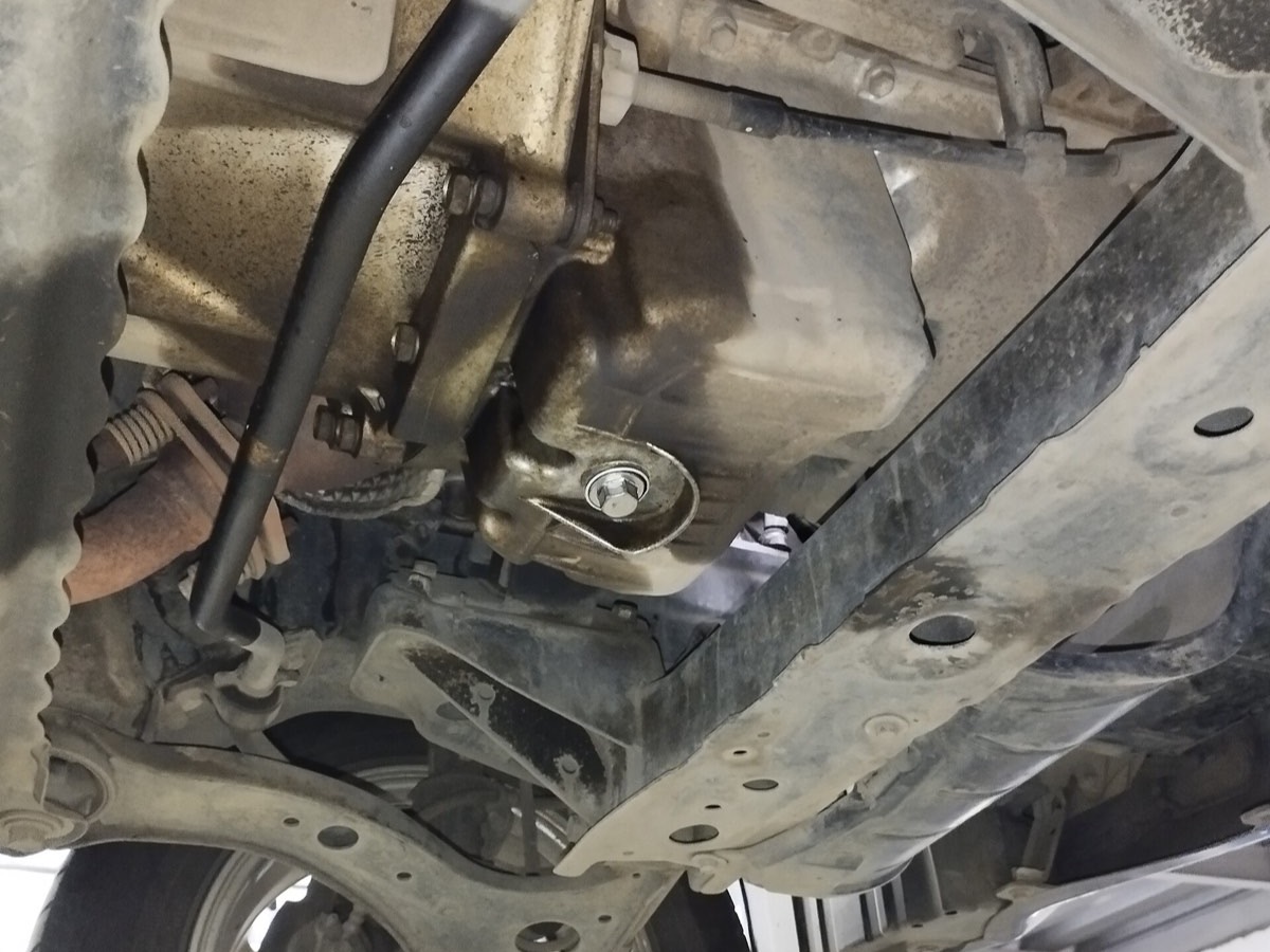 A car on a workshop lift from below, the oil pan and drain plug sitting just above a subframe crossmember