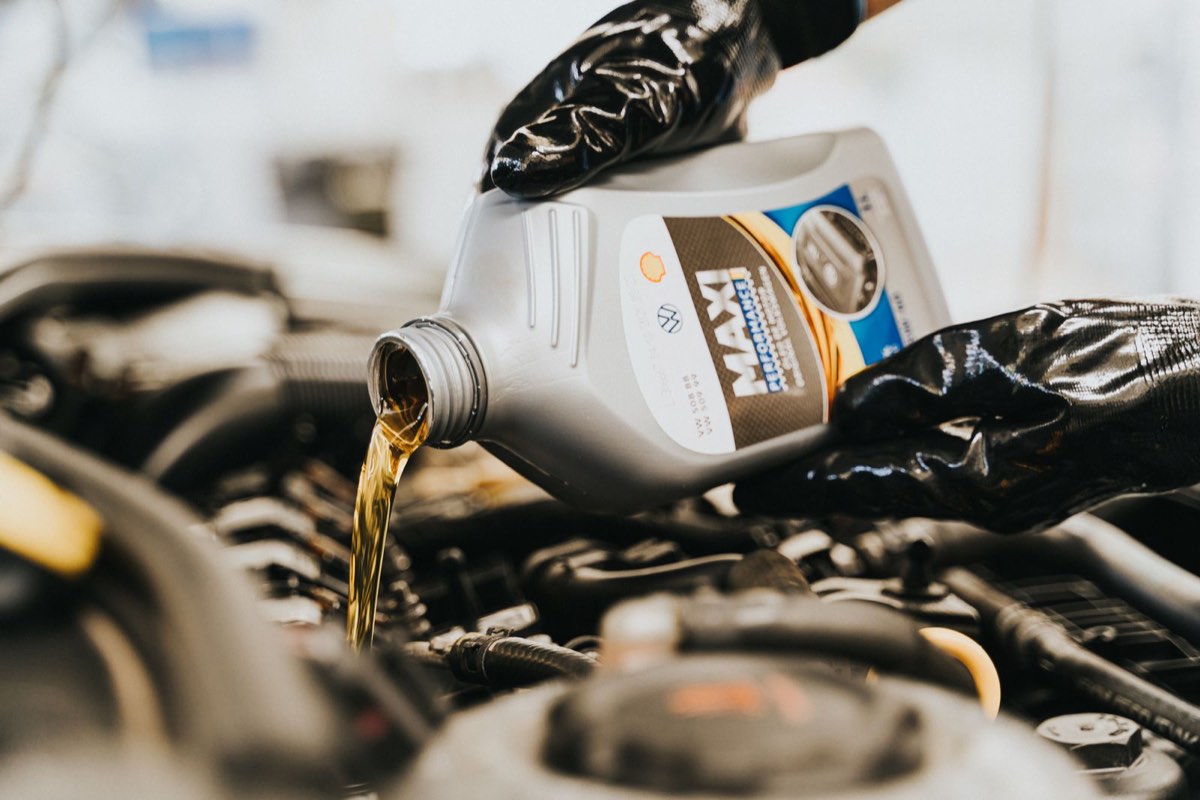 Gloved hands pouring engine oil from a plastic bottle into an engine filler neck