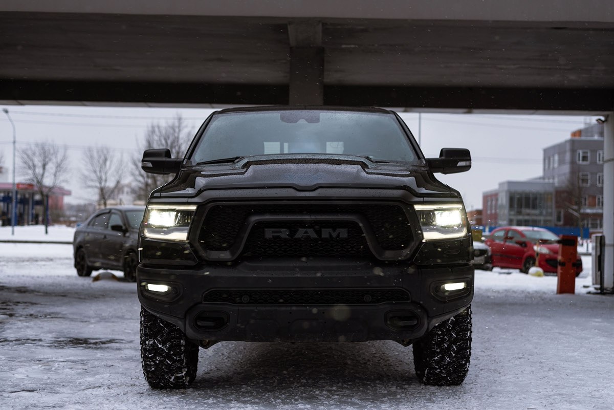 Black Ram 1500 pickup seen head-on under a bridge on a snow-covered street, headlights on