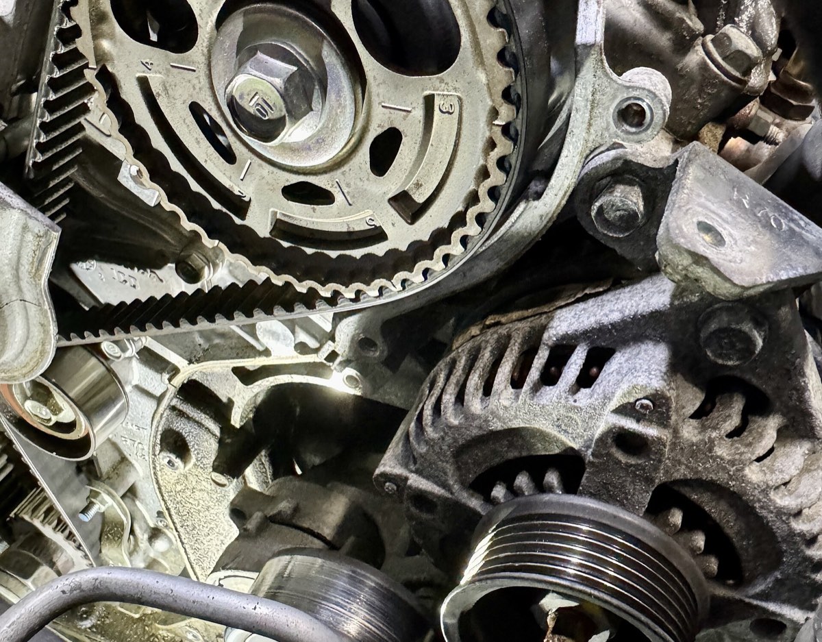 A toothed rubber timing belt wrapped around a camshaft sprocket, with the tensioner and alternator visible beside it