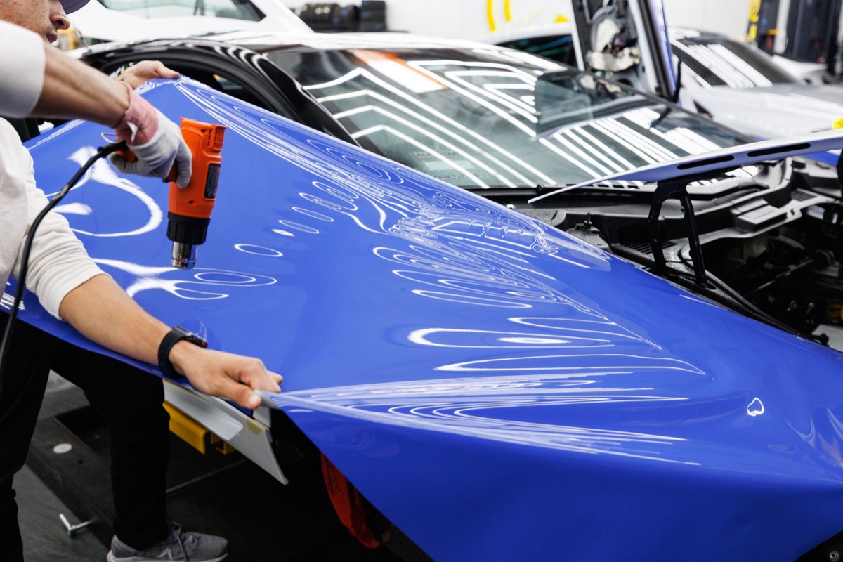 A technician using a heat gun and a squeegee to lay blue vinyl film over a car body panel in a workshop